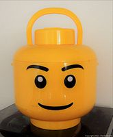 Front view of the yellow Lego robot head carrying case with smiling face and handle on top.