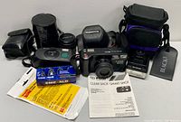 Wide shot of all items including Fuji camera with manual, Canon Zoom camera, Olympus lens case, flash, cases, and cleaning film.