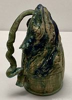 Side view of the pottery vessel shaped like a chameleon or iguana head with twisted handle and textured blue-green glaze.