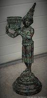 Front angle view of bronze statue showing young male figure in wizard or jester costume holding basket candleholder with woven details.
