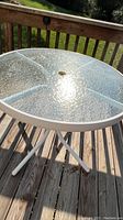 Round outdoor patio table showing textured glass top with central umbrella hole and white aluminum frame on a wooden deck in sunlight.