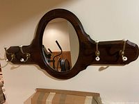 Full front view of the colonial-style dark stained pine mirror with oval glass and five brass coat hooks with ceramic knobs.