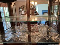 Glass cabinet shelf holding multiple clear Fenton hobnail glass items including bowls, compotes, and trays with hobnail patterns.