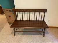 Front view of dark wooden bench with spindle backrest and carved floral detail on top rail.