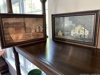 Two framed farmhouse pictures on a dark wood furniture surface near window light. Both have wood frames and depict muted rustic scenes.