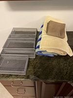 Five metal mesh drawer inserts arranged in a stack and three towels next to them on a countertop: cream towel with a brown washcloth, and blue and white striped towel.