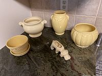 Five pieces of Shawnee pottery displayed on granite countertop: three vases, one floral pot with saucer, and a cow-shaped planter.