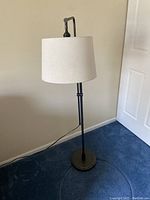 Full view of the black metal floor lamp with white fabric drum shade showing its base and cord on blue carpet.