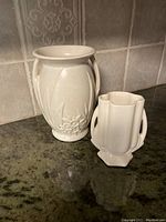 Two vintage off-white pottery vases displayed on countertop with tiled backsplash. The taller vase has floral relief and two handles; the smaller is hexagonal with dual handles.