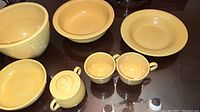 Front view of seven yellow Fiesta Ware pottery items including bowls, sugar bowl with lid, and coffee cups with handles.