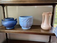 Three vintage pottery pieces on shelf including blue Shawnee rounded vase with scroll handles, light blue McCoy planter with leaf pattern, and peach Shawnee vase with vertical ribbing and small handles.
