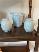Three vintage pottery pieces displayed on a wooden shelf: a white glazed pitcher with leaf embossing and two white lidded jars with horizontal ridges and leaf patterns.