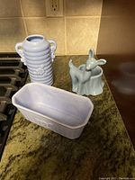 Three pottery pieces on countertop showing ribbed vase with handles, rectangular planter, and deer figurine in pastel blue glaze