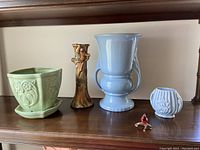 All five items in the lot arranged on a wooden shelf: green Shawnee planter with owl pattern and saucer, McCoy tree trunk vase with fruit motifs, large pale blue vase, small white ribbed floral vase, and small cardinal bird figurine.
