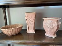 Three vintage mauve pink Shawnee pottery pieces on wood shelf: oval bowl, rectangular vase, square floral relief vase.