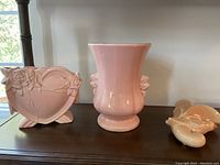 Three McCoy pottery pieces on a wooden surface: heart-shaped floral vase, urn-shaped rose vase, bird wall pocket.