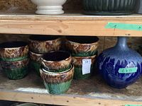 Wide shelf view showing nine brown-green drip glaze planter pots of varying heights alongside one spherical blue ceramic vase with white speckle glaze.