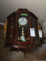 Front view of wooden wind up wall clock with glass door showing clock face and pendulum, includes key.