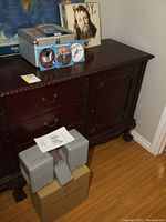 Metal case on dark wood sideboard with six vintage LPs behind it and wireless speakers placed in front near boxes.