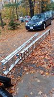 Two aluminum ladders side by side on leaf-covered ground outdoors with a car in the background. The ladders are laid flat on the ground.