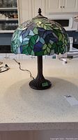 Full view of the lamp showing stained glass shade with leaf pattern and tree trunk base on a white countertop, lamp is unplugged.