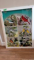 Full view of the framed painted glass panel showing six bird scenes, with teal border and white frame on a wood floor.