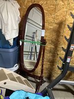 Full oval mirror standing on wooden frame with turned vertical supports and curved feet, showing entire mirror with elastic bands securing it.