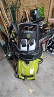 Front view showing green Sun Joe pressure washer with power cord wrapped around front and two white detergent bottles.