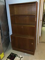 Front view of the teak secretary cabinet showing open upper shelving and lower cabinet with sliding doors and recessed handles.