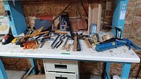 View of assorted hand tools and power driver arranged on white workbench surface with tool belt behind. Metal tools have wooden and plastic handles.