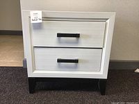 Front view of a white bedside table showing two drawers with black handles and short legs.