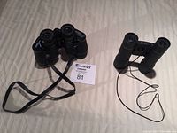 Two pairs of binoculars side by side displayed on beige surface, one with neck strap and the other with lanyard