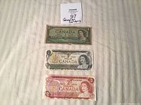 Front view of three Canadian banknotes: 1954 one dollar, 1973 one dollar, and 1974 two dollar bills, displayed on a fabric surface.