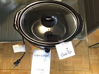 Oval black Crock-Pot slow cooker with glass lid and instruction manual on a glass surface. Power cord and plug visible, control knob in front.
