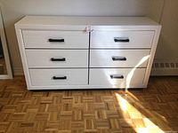 White and light grey wooden dresser with six drawers and black handles, photographed in natural light on parquet flooring.