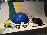Blue Bosu-style balance ball with black base and attached resistance bands, foot pump, green and yellow therabands, black balance disk, storage bag, and red dodgeball.