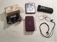 Photo showing all items laid out: opera glasses with pouch, cigarette case, pince-nez glasses with case and cord, red box with crown emblem.