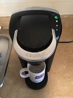 Top view of the Keurig coffee maker on a counter with a white mug underneath, machine powered on and ready to brew.