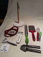 Lot contents showing all utensils arranged on a light fabric background: paper towel rack, apple slicer, can opener, graters, whisks, garlic press, and peeler.