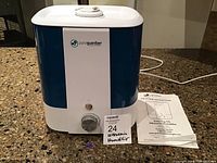 Ultrasonic humidifier with white top and base, blue translucent water container, control knob, and instruction manual and cleaning brush laid beside it on a countertop, lit by room light.