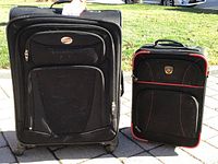Front view of large black American Tourister suitcase with multiple zippered pockets and smaller black carry-on with red piping in sunlight
