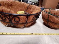 Two black wire hanging baskets with brown coconut liners, side by side with measuring tape showing sizes.
