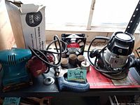 Lot of vintage power and hand tools placed on a black tabletop under a window including Craftsman router, Makita sander, sanding papers, sanding discs, several cylindrical sanding paper rolls, and a hack saw frame.