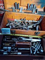 Orange Craftsman toolbox open showing assorted sockets, wrenches, and accessories inside the two tray sections. Visible surface wear and grime.