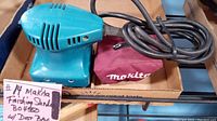 Photo showing overall Makita finishing sander with power cord and attached dust bag inside a cardboard box.