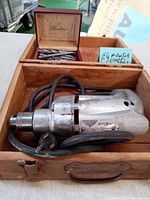 Electric drill placed inside an open wooden box showing the drill, cord, and plug with a visible Miller Falls label.