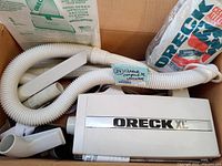 Oreck XL Compact vacuum canister with attached hose, assorted attachments and vacuum bag pack inside a cardboard box.