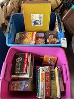 Two storage bins containing various books and puzzles. The bin in the back is blue and holds The Chronicles of Narnia, Nickelodeon Magazine, and other books and puzzles. The front bin is pink and holds several books including some on bodybuilding, history, and puzzles.
