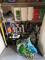 View of three shelves filled with assorted cleaning supplies including sprays, wipe containers, and paper towels, with cleaning cloths and larger bottles on the floor area beneath the shelves.
