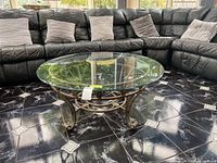 Photo showing glass top coffee table placed in front of a black sectional sofa with pillows on a tiled floor.